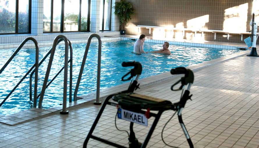 I förgrunden en rollator med en skylt på med namnet Mikael. I bakgrunden badar två personer i en pool.
