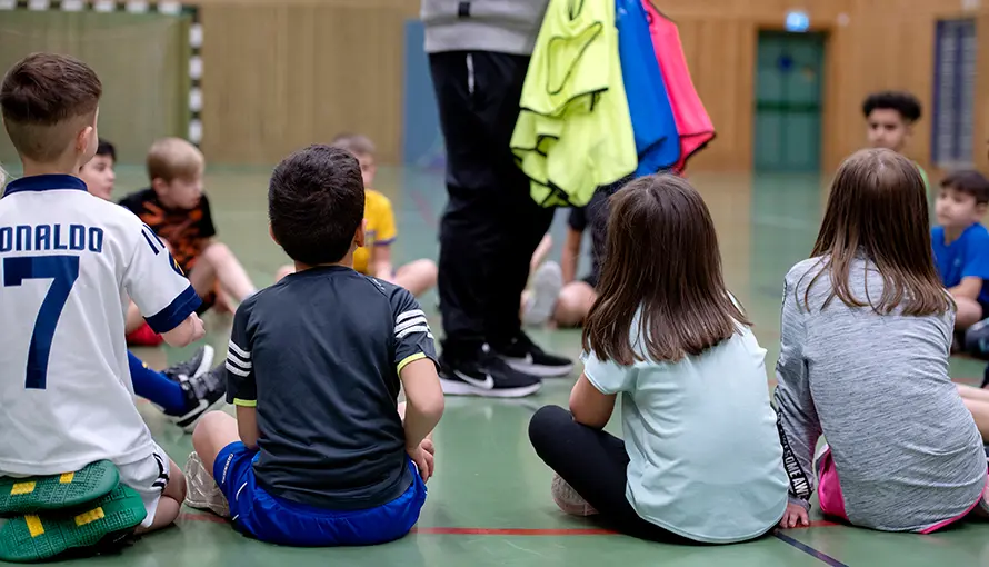 Barn sitter på golv i gymnastiksal och lyssnar på ledaren. 