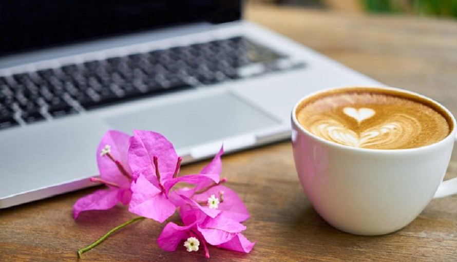 En laptop står på ett bord. Bredvid datorn står en kaffekopp samt att det ligger en rosa blomma vid datorn.