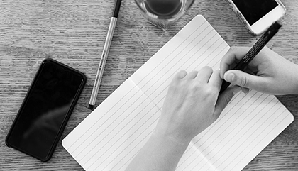 En persons händer som håller i en penna över ett skrivbord med anteckningsblock och mobil.