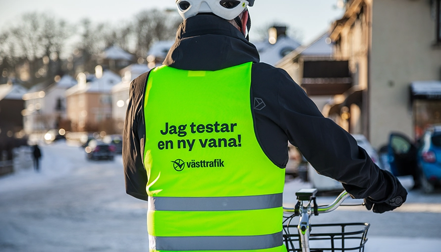 En person bär en neon gul väst med texten ”Jag testar en ny vana!” och logotypen ”västtrafik”. Personen står bredvid en cykel på snöig bakgrund.
