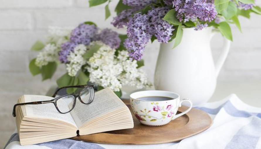 På ett bord med syrener ligger en bok, ett par glasögon och en kopp kaffe.