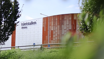 "Iskristallen Arena" syns på en byggnad med en fasad i rostrött och silver. En väg och grön vegetation finns i förgrunden.