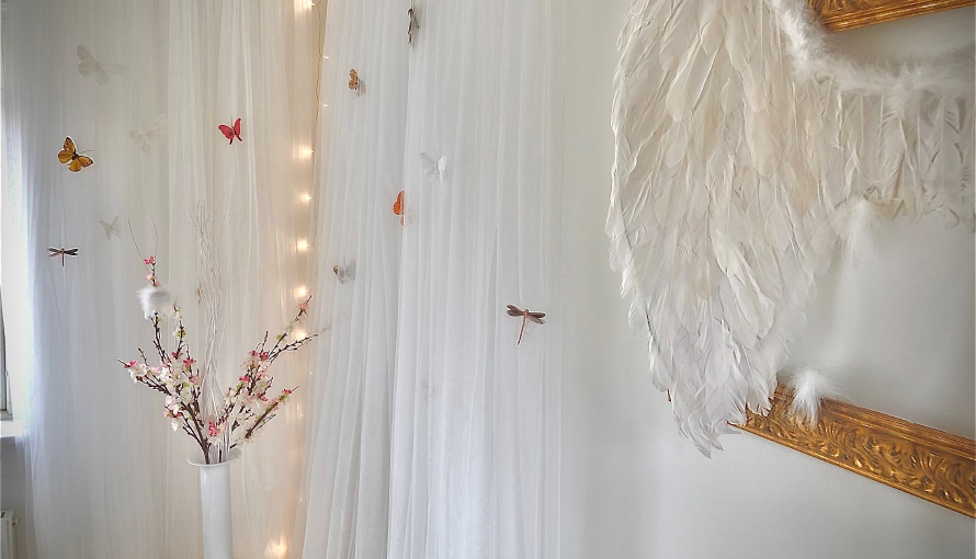 Ett rum med vita väggar och vita gardiner, konstgjorda fjärilar i gardinen. Änglavingar hänger på en guldram.