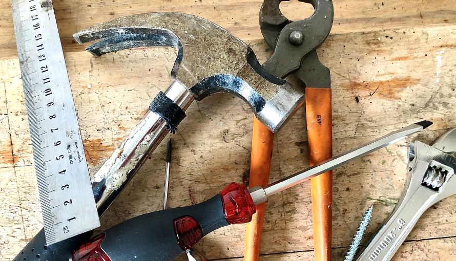 En hammare, en skiftnyckel, en tång, en skruvmejsel och ett måttband ligger på ett träbord.