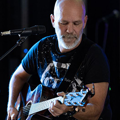 Artisten Rocktrubaduren sitter bakom en mikrofon på en scen, hållandes i en akustisk gitarr