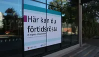 "Här kan du förhandsrösta" 26 augusti - 13 september. En affisch i ett fönster med en byggnad och träd i bakgrunden.