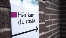 "Här kan du rösta" och en pil som pekar åt höger på en vit skylt mot en tegelvägg.