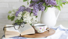 På ett bord med syrener ligger en bok, ett par glasögon och en kopp kaffe.