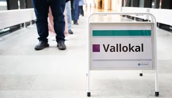“Vallokal” står på ett metallstöd i en korridor, med personer i bakgrunden.
