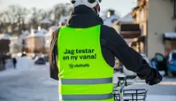 En person bär en neongrön reflexväst med texten "Jag testar en ny vana!" och logotypen "västtrafik". Personen står bredvid en cykel på en snöig gata.