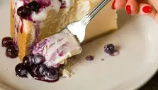En bit cheesecake med blåbär och en gaffel. Blåbärssås syns runt kanten på tallriken.